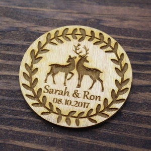 May include: A round wooden wedding favour with a wreath design and two deer silhouettes. The favour is personalised with the names "Sarah & Ron" and the date "08.10.2017".