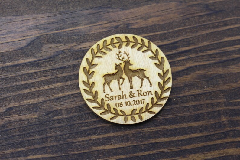 May include: Wooden round ornament featuring a wreath design and two deer silhouettes. The ornament is engraved with the text "Sarah & Ron 08.10.2017".