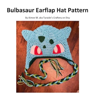 Puede incluir: Un gorro de Bulbasaur de crochet azul con lazos trenzados verdes y azules. El gorro tiene ojos de fieltro rojo y una nariz de fieltro verde. El gorro está sobre una superficie de madera. Dificultad: Fácil.
