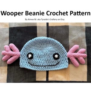 May include: A light blue crochet beanie with pink side fins and a black smiling face. The text "Wooper Beanie Crochet Pattern" is at the top, with the designer's name below. The beanie is handmade and likely for a child.