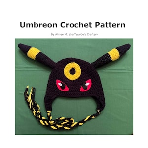 Pode incluir: Chapéu preto de crochê com detalhes amarelos e vermelhos, inspirado em um personagem de uma franquia popular. O chapéu apresenta orelhas grandes com listras amarelas, olhos vermelhos e um círculo amarelo na testa. O texto "Umbreon Crochet Pattern" está no topo.