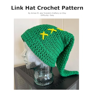 Puede incluir: Un gorro verde tejido a crochet con detalles de cruces amarillas, inspirado en el personaje de Link. El gorro tiene un extremo largo y puntiagudo. El texto de la imagen dice "Link Hat Crochet Pattern" y "Difficulty: Easy".