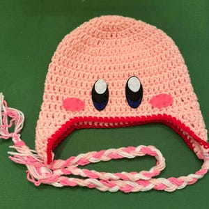 Pode incluir: Um chap&eacute;u rosa de croch&ecirc; feito &agrave; m&atilde;o com um design de personagem de desenho animado. O chap&eacute;u apresenta olhos pretos e brancos, bochechas rosa e uma borda vermelha. Fios tran&ccedil;ados rosa e branco se estendem do chap&eacute;u.