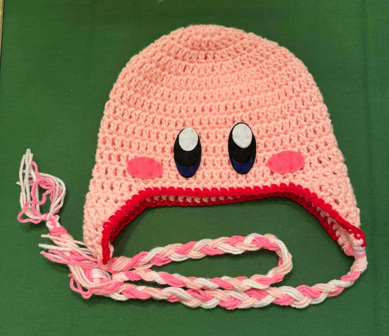 Pode incluir: Um chap&eacute;u rosa de croch&ecirc; feito &agrave; m&atilde;o com um design de personagem de desenho animado. O chap&eacute;u apresenta olhos pretos e brancos, bochechas rosa e uma borda vermelha. Fios tran&ccedil;ados rosa e branco se estendem do chap&eacute;u.