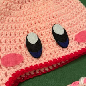 Pode incluir: Um chap&eacute;u rosa de croch&ecirc; feito &agrave; m&atilde;o com um design de personagem de desenho animado. O chap&eacute;u apresenta olhos pretos e azuis, bochechas rosa e uma borda vermelha. O chap&eacute;u &eacute; feito de fio macio e &eacute; adequado para crian&ccedil;as.