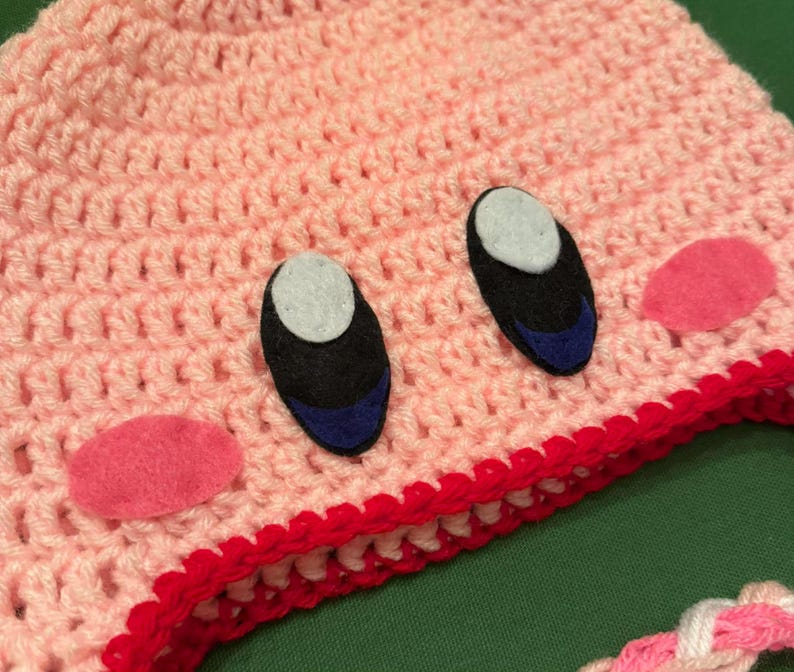 Pode incluir: Um chap&eacute;u rosa de croch&ecirc; feito &agrave; m&atilde;o com um design de personagem de desenho animado. O chap&eacute;u apresenta olhos pretos e azuis, bochechas rosa e uma borda vermelha. O chap&eacute;u &eacute; feito de fio macio e &eacute; adequado para crian&ccedil;as.