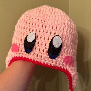 Pode incluir: Um chap&eacute;u rosa de croch&ecirc; com um design de personagem de desenho animado. O chap&eacute;u apresenta olhos grandes em preto e branco com detalhes em azul, bochechas rosa e uma borda vermelha. O chap&eacute;u &eacute; feito &agrave; m&atilde;o e parece ser um acess&oacute;rio de inverno.