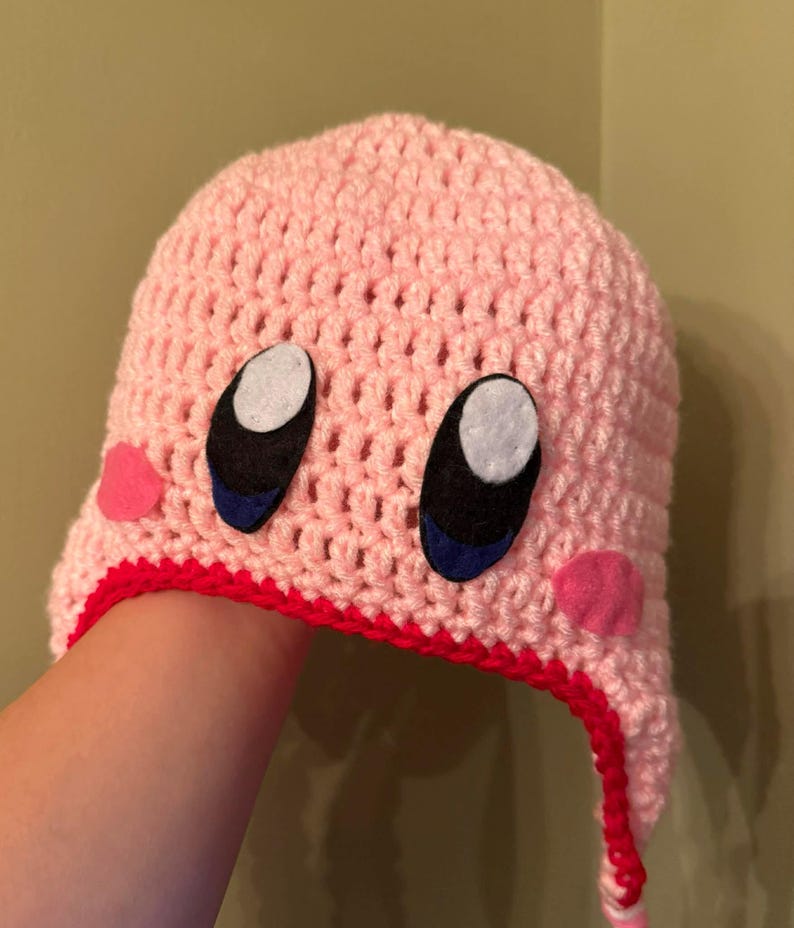 Pode incluir: Um chap&eacute;u rosa de croch&ecirc; com um design de personagem de desenho animado. O chap&eacute;u apresenta olhos grandes em preto e branco com detalhes em azul, bochechas rosa e uma borda vermelha. O chap&eacute;u &eacute; feito &agrave; m&atilde;o e parece ser um acess&oacute;rio de inverno.