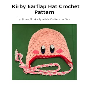 Pode incluir: Um chap&eacute;u rosa de croch&ecirc; com abas para as orelhas, acabamento vermelho e la&ccedil;os tran&ccedil;ados. O chap&eacute;u apresenta olhos bordados e bochechas rosa, que se assemelham a um personagem popular de videogame. O texto "Kirby Earflap Hat Crochet Pattern" est&aacute; no topo.