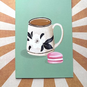 May include: A rectangular greeting card featuring a printed illustration of a speckled ceramic mug filled with brown coffee, decorated with a large white flower and dark leaves, positioned next to a single pink macaron on a solid mint green background.