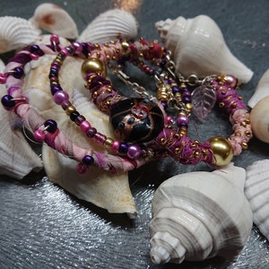 Op de afbeelding: Een set van drie armbanden gemaakt met roze, paarse en gouden stof, kralen en een zwarte glazen bedel. De armbanden worden getoond op een bed van witte schelpen.