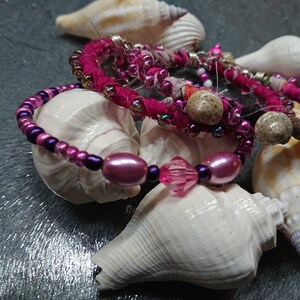 May include: A set of three bracelets with pink, purple, and white beads, wrapped around seashells.
