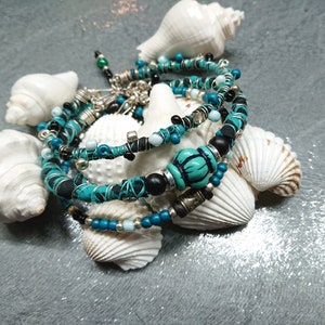 Op de afbeelding: Een set van vier turquoise en zwarte kralen armbanden met zilveren accenten, getoond op een bed van witte schelpen.