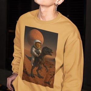 May include: Mustard yellow sweatshirt with a crew neck and long sleeves. The graphic print depicts a cat in an astronaut suit riding a dinosaur on a Mars-like planet. The background features a large red planet. The sweatshirt is made of a soft material.