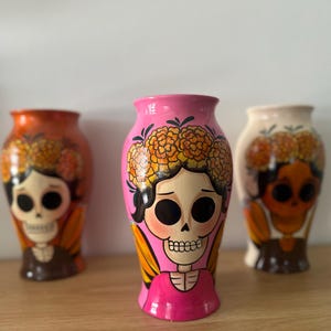 May include: Three ceramic vases featuring hand-painted skull figures with floral crowns. The central vase is bright pink, while the flanking vases are orange and cream. Each figure displays large black eye sockets, a skeletal nose, and teeth.