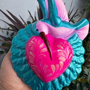 May include: A hand-painted, heart-shaped wall hanging featuring a hummingbird design. The sculpture has a vibrant color palette of pink, teal, and gold. The hummingbird's beak is a dark brown, and the heart has a decorative pattern.
