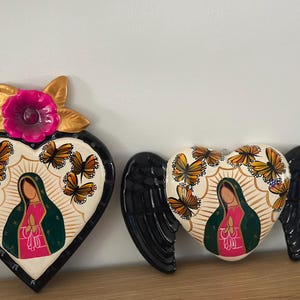 May include: Two heart-shaped wall plaques featuring the Virgin of Guadalupe. The left plaque has a pink flower at the top and orange butterflies. The right plaque features black wings on the sides and orange butterflies surrounding the central figure.