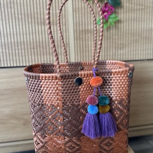 May include: A woven tote bag featuring a geometric pattern in shades of brown and copper. The bag has two braided handles and a decorative charm consisting of multi-colored pom-poms and two purple tassels attached to the front rim with a metal snap.