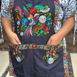 May include: A dark gray apron with a vibrant embroidered hummingbird design. The apron features colorful floral accents and a decorative border. It has two pockets and a neck strap.