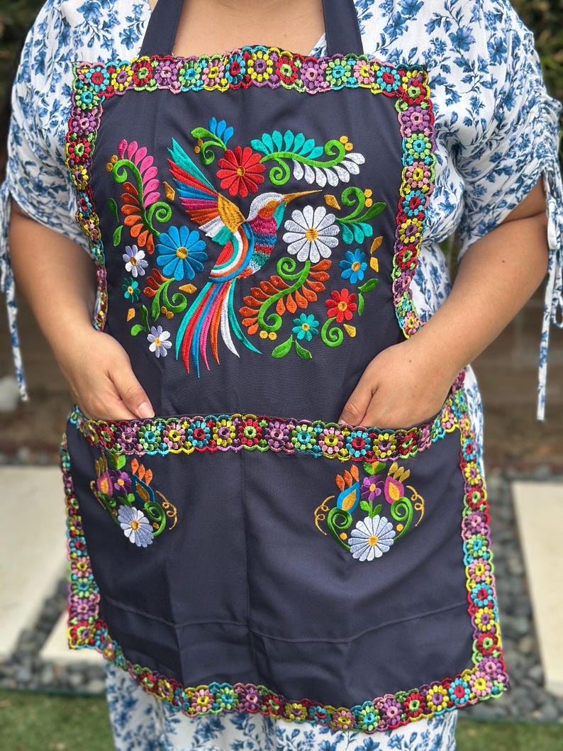 May include: A dark gray apron with a vibrant embroidered hummingbird design. The apron features colorful floral accents and a decorative border. It has two pockets and a neck strap.