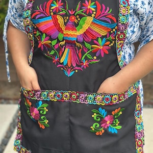 May include: Black apron with a vibrant, embroidered design featuring a colourful bird with outstretched wings, surrounded by flowers. The apron has two pockets and is trimmed with a multi-coloured floral border.