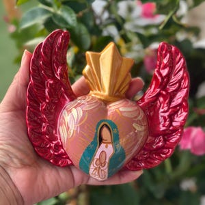 May include: A heart-shaped religious figurine with red wings and a gold crown. The heart is painted pink with a depiction of the Virgin Mary in blue and gold. The figurine is held in a hand.