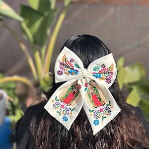 May include: A white fabric bow with colorful embroidery featuring a religious image and floral designs. The bow is tied in a large, symmetrical shape.