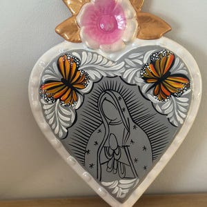 May include: A heart-shaped wall plaque featuring a central gray illustration of a figure in a hooded robe with hands clasped in prayer. Two orange and black monarch butterflies are painted on the sides, topped with a pink flower and gold leaves.