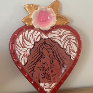 May include: A heart-shaped religious wall hanging with a red border and a central image of a religious figure. The image is surrounded by white decorative elements. A pink and white flower with gold leaves adorns the top.