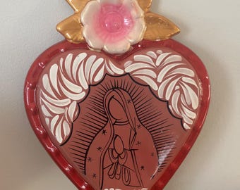 May include: A heart-shaped religious wall hanging with a red border and a central image of a religious figure. The image is surrounded by white decorative elements. A pink and white flower with gold leaves adorns the top.