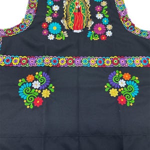 May include: Black apron with colorful floral embroidery and trim. The apron features a central embroidered image of the Virgin Mary, surrounded by flowers in shades of pink, blue, orange, and white. The edges are trimmed with a multi-colored crochet design.