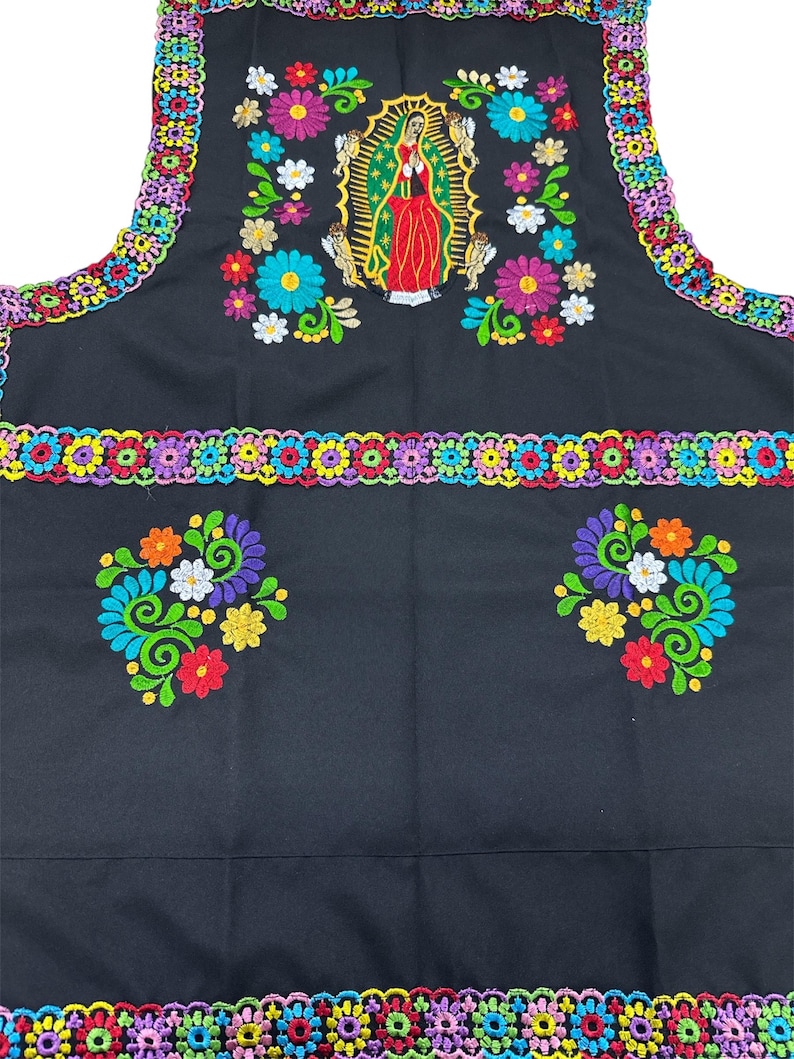 May include: Black apron with colorful floral embroidery and trim. The apron features a central embroidered image of the Virgin Mary, surrounded by flowers in shades of pink, blue, orange, and white. The edges are trimmed with a multi-colored crochet design.