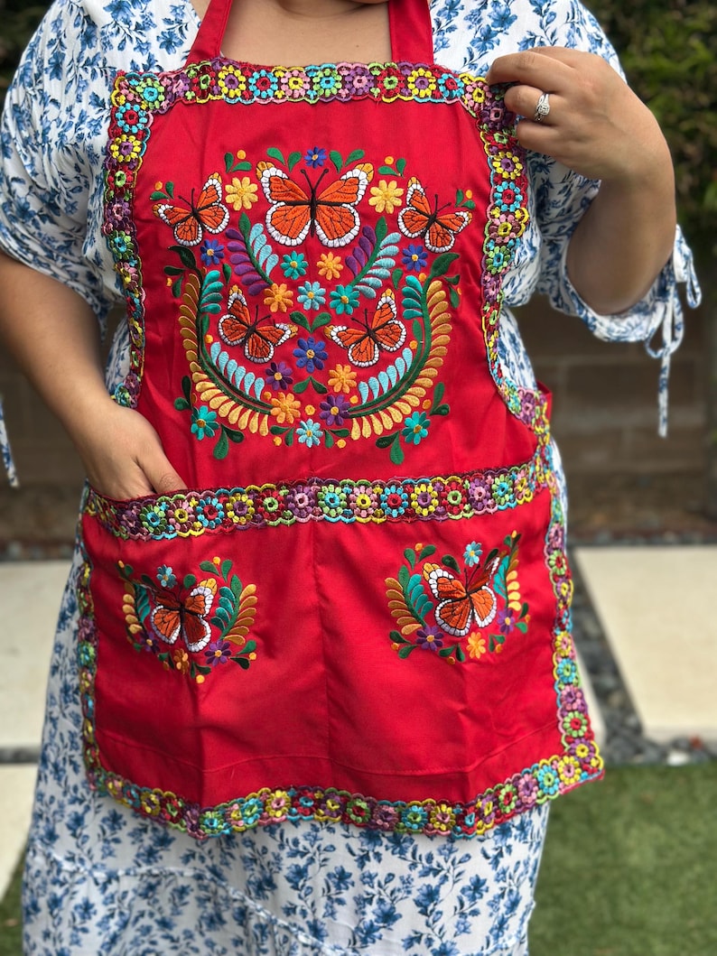 Pu&ograve; includere: Un grembiule rosso acceso con ricami colorati di farfalle e motivi floreali. Il grembiule presenta due tasche e bordi decorativi. Il ricamo delle farfalle include dettagli arancioni, gialli, blu e verdi.