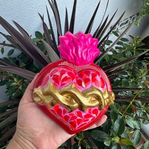 May include: A vibrant, heart-shaped decorative piece with a glossy red finish. It features a gold-colored crown of thorns and a pink flame at the top. The heart is filled with red and pink heart-shaped patterns.