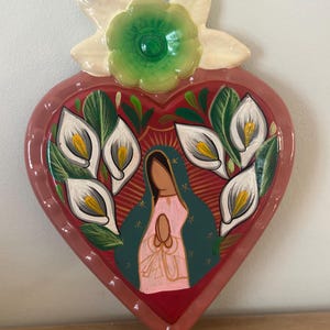 May include: A heart-shaped ceramic wall hanging with a religious theme. The design features a central image of a figure in prayer, surrounded by white calla lilies and green foliage, set against a red background. A green flower and white petals adorn the top.