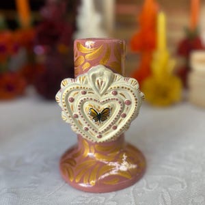 May include: A pink ceramic candle holder with gold leaf accents and a heart-shaped embellishment. The heart features a butterfly and is adorned with small pink dots. The candle holder has a flared base and a cylindrical top.