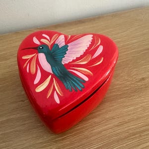 May include: A heart-shaped, red wooden box with a painted hummingbird design. The hummingbird is teal and pink, with a long beak and is surrounded by floral accents. The box is closed and sits on a wooden surface.