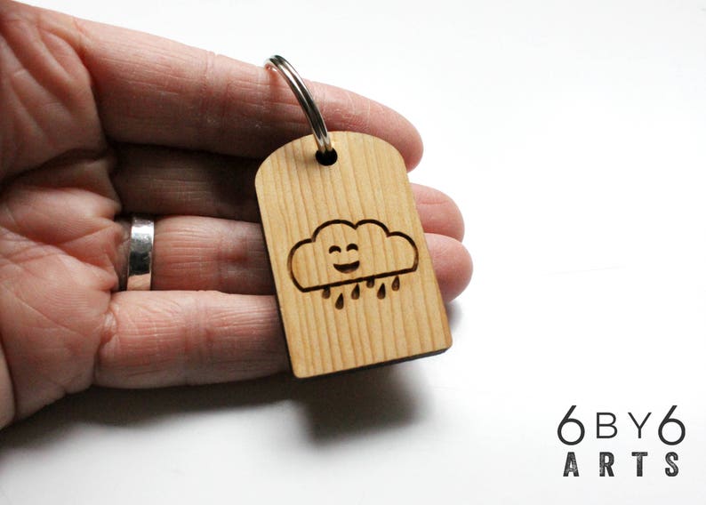 May include: Wooden keychain with a smiling cloud and raindrops design. The keychain is made of light-colored wood and has a silver-colored metal ring. The text "6by6 ARTS" is visible in the bottom right corner.