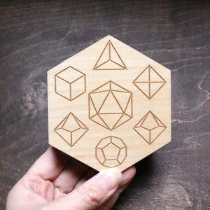 Peut inclure: Un dessous de verre en bois clair de forme hexagonale. Le dessous de verre présente des formes géométriques gravées, dont un cube, une pyramide et un dodécaèdre. Le dessous de verre est conçu pour les boissons et est un objet décoratif.