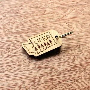 May include: A wooden keychain with a laser-etched design. The keychain is shaped like the state of Washington, with the word "LIFER" and a row of trees etched inside. It has a silver-colored metal keyring.