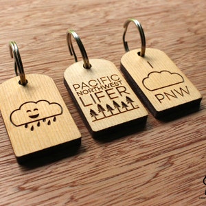May include: Three wooden keychains with metal rings. One keychain features a smiling cloud with raindrops, another reads "PACIFIC NORTHWEST LIFER" with trees, and the last says "PNW" with a cloud.
