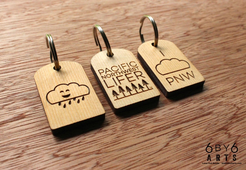 May include: Three wooden keychains with metal rings. One keychain features a smiling cloud with raindrops, another reads "PACIFIC NORTHWEST LIFER" with trees, and the last says "PNW" with a cloud.