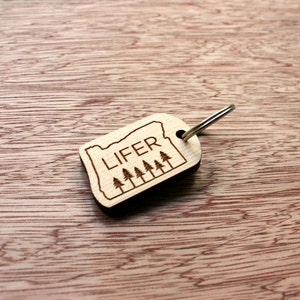 May include: A wooden keychain featuring the outline of Oregon with the word "LIFER" and a row of trees engraved on it. The keychain has a silver metal ring attached. The background is a wooden surface.