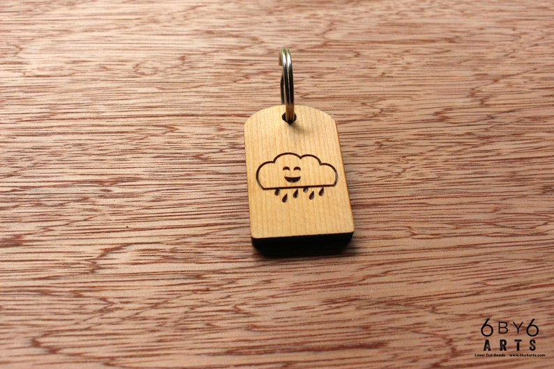May include: Wooden keychain with a smiling cloud and rain design. The keychain is made of light-colored wood and has a silver-colored metal ring. The background is a wooden surface. The text "6by6 arts" is visible.