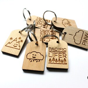 May include: Wooden keychains with engraved designs, including mountains, clouds, and trees. Some keychains feature the text "PACIFIC NORTHWEST LIFER" and "PNW". Each keychain has a silver metal ring.