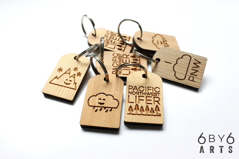 May include: Wooden keychains with engraved designs, including mountains, clouds, and trees. Some keychains feature the text "PACIFIC NORTHWEST LIFER" and "PNW". Each keychain has a silver metal ring.
