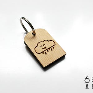 May include: Wooden keychain with a smiling cloud and raindrops design. The keychain is rectangular with rounded top corners and a silver metal ring. The cloud and raindrops are etched into the light-colored wood.