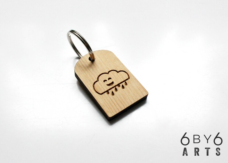 May include: Wooden keychain with a smiling cloud and raindrops design. The keychain is rectangular with rounded top corners and a silver metal ring. The cloud and raindrops are etched into the light-colored wood.