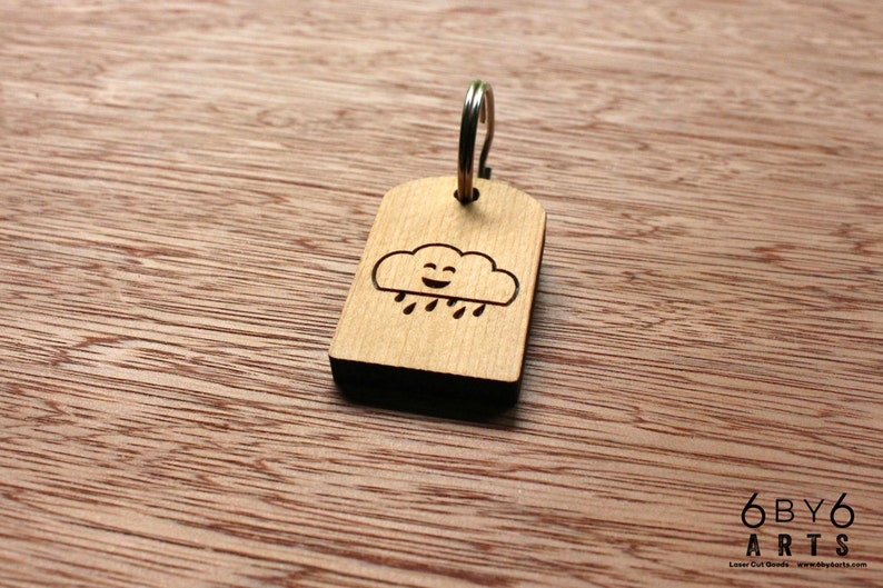 May include: Wooden keychain with a smiling cloud and rain design. The tag is rectangular with rounded corners and a silver key ring. The keychain is made of light-colored wood.