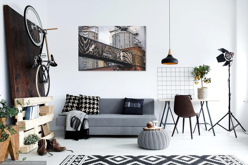 May include: A modern living room interior with a gray sofa, black and white patterned pillows, a black throw blanket, a black and white rug, a gray knitted ottoman, a black pendant light, a black and white grid wall decor, a wooden table with a brown chair, a potted citrus tree, a black studio light, and a bicycle hanging on a wooden wall.
